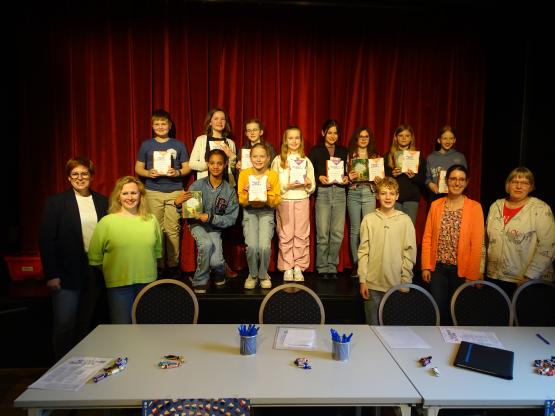 Im Vordergrund: Jurymitglieder (von links) Rabea Klaas (Buch und Kunst Blanke), Christina Schürmann (Filialleiterin Thalia Lingen), Mika Ekelhoff (Vorjahressieger des bundesweiten Vorlesewettbewerbs), Juliane Schmalisch-Fischer (Leiterin der Trinitatis-Bücherei in Lingen) und Mechthild Jasper (Stadtbibliothek Lingen). Im Hintergrund auf der Bühne (von links nach rechts): Vorleser*innen Ole Zimmermann, Jule Behner (hinten), Naemi She-Ra Sieverding (vorne), Luisa Poppen (hinten), Merle Winter (vor