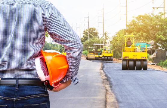 Ingenieur, der Schutzhelm an der Straßenbaustelle mit Rollenverdichterarbeitsasphaltstraße hält